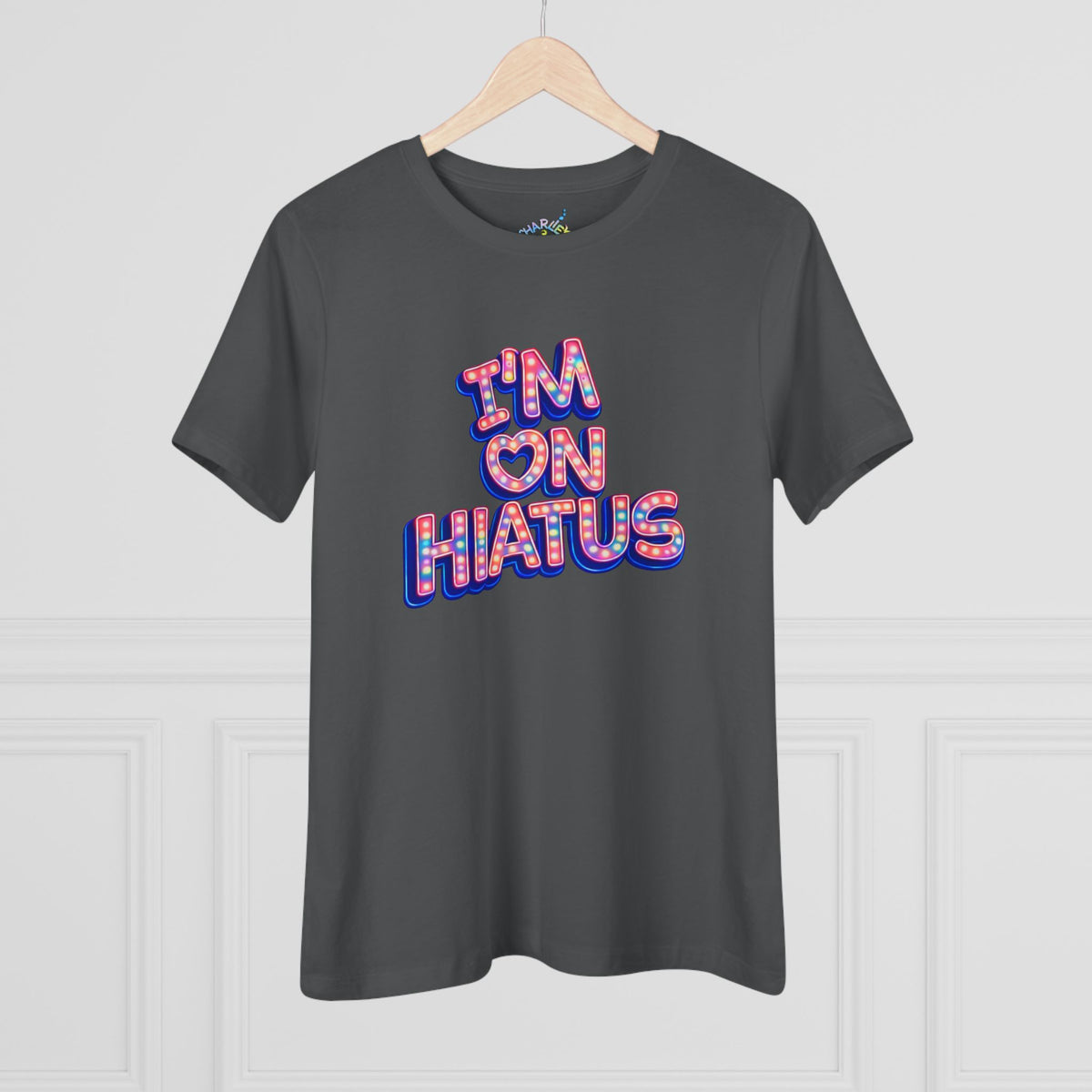 A dark gray crewneck t-shirt hanging on a wooden hanger against a white paneled wall. The shirt features a graphic text print reading 'I'M ON HIATUS' in a 3D bubble font filled with a red, white, pink, and blue pattern.