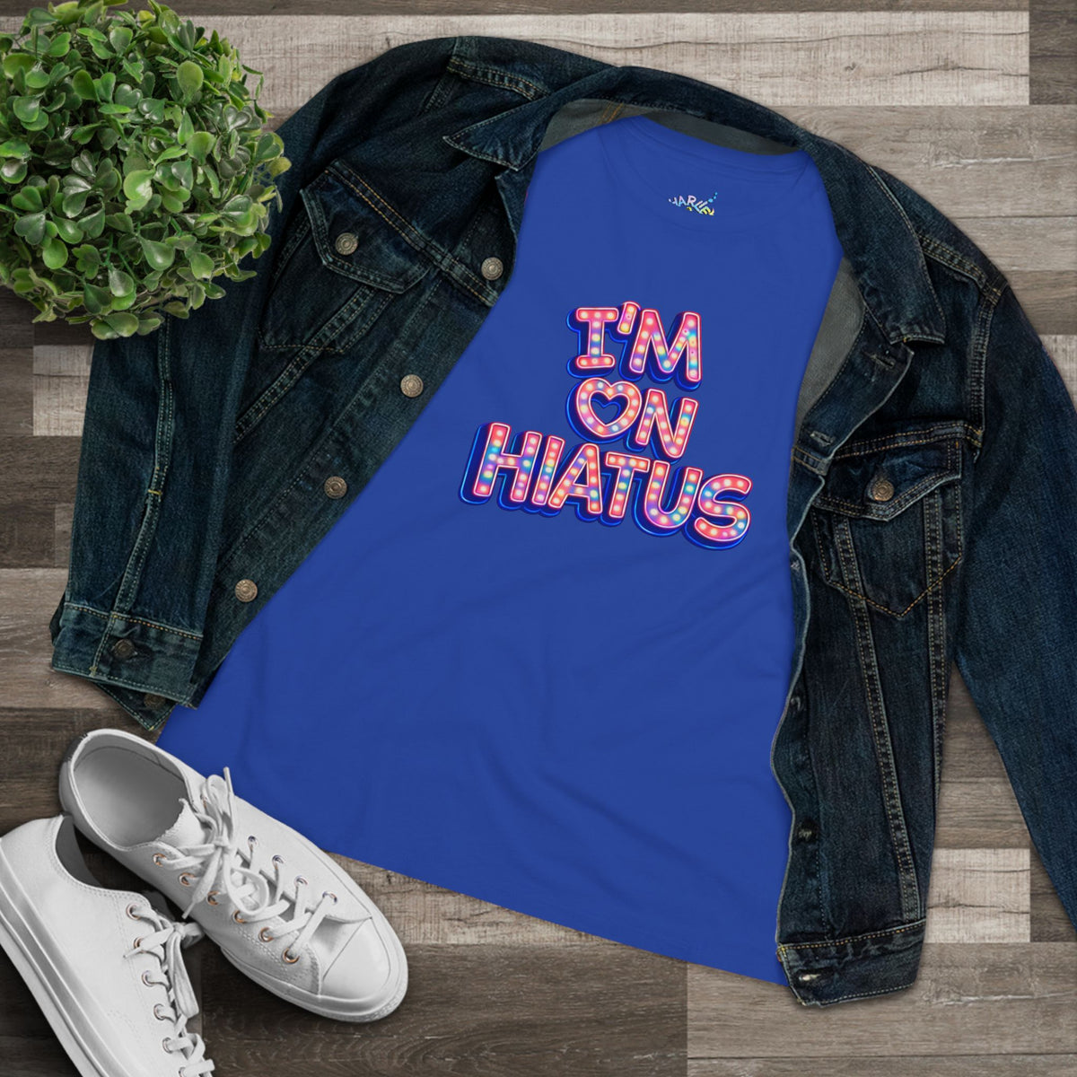 A royal blue tank top with a colorful retro-style graphic reading 'I'M ON HIATUS' with a heart, styled in a flat lay with a dark denim jacket, white sneakers, and a small green plant on a wood plank background.